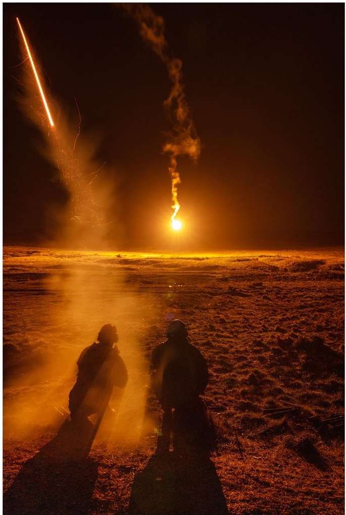 Two people stand silhouetted in an orange-lit landscape as bright light and smoke plumes fill the horizon at night in a wide shot.