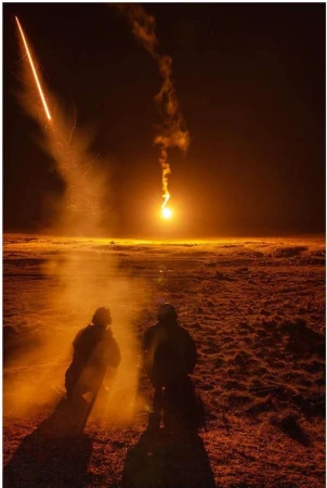 Two people stand silhouetted in an orange-lit landscape as bright light and smoke plumes fill the horizon at night in a wide shot.