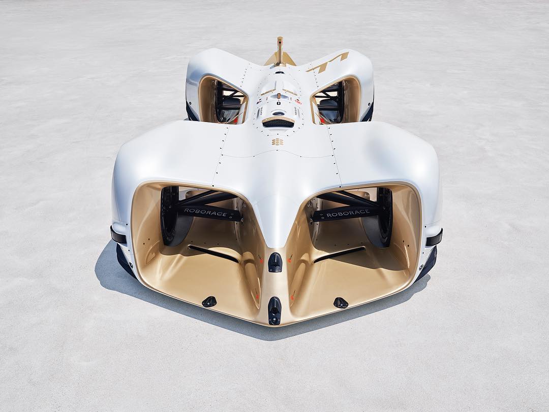 A futuristic white and gold race car is shown up close from above on a flat, light grey outdoor surface on a sunny day.