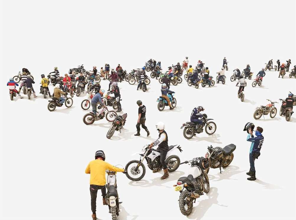 Wide shot of many people riding vintage motorcycles on a white surface, with some people standing nearby, creating a sense of community.