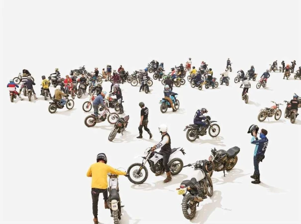 Wide shot of many people riding vintage motorcycles on a white surface, with some people standing nearby, creating a sense of community.