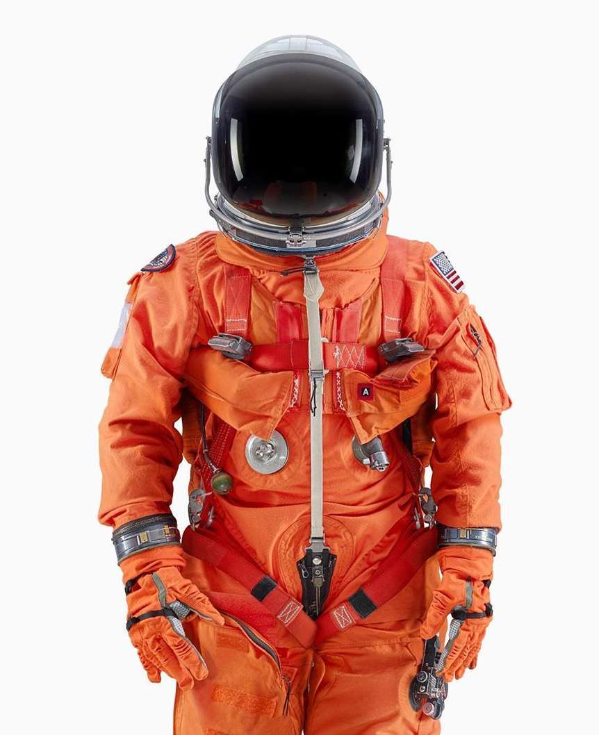 An astronaut in a bright orange spacesuit and helmet stands in front of a white background in a studio shot, space exploration theme.