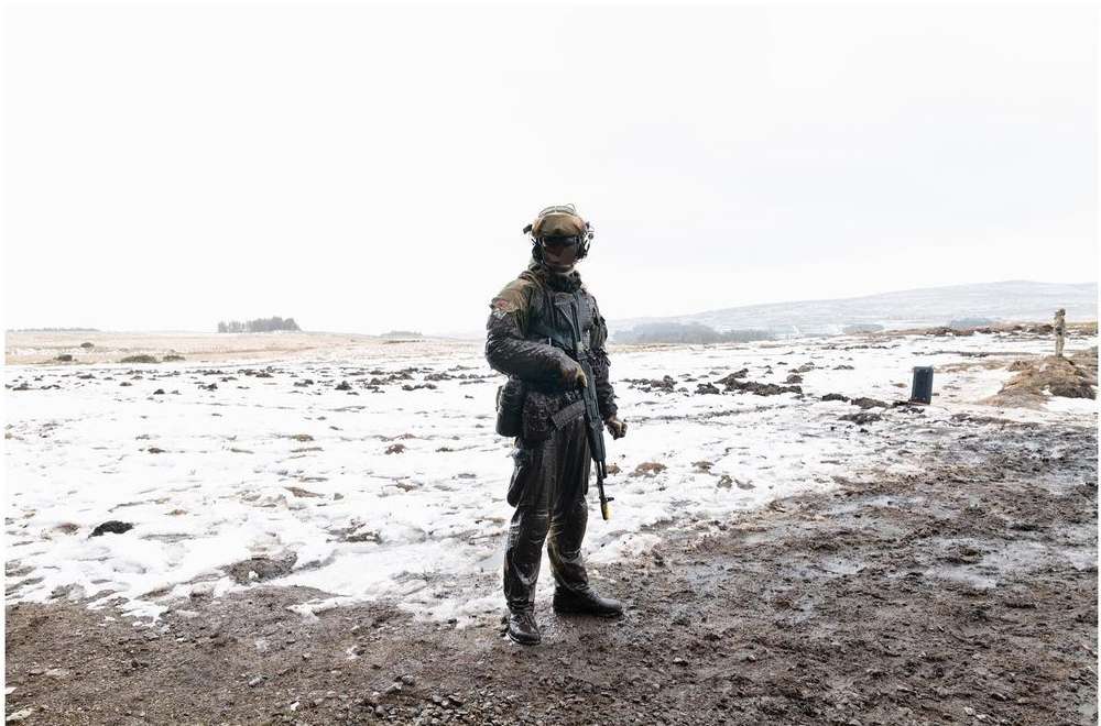 Full-body shot of a soldier in tactical gear holding a rifle in a snowy, rural landscape under an overcast sky, depicting a scene of military readiness.