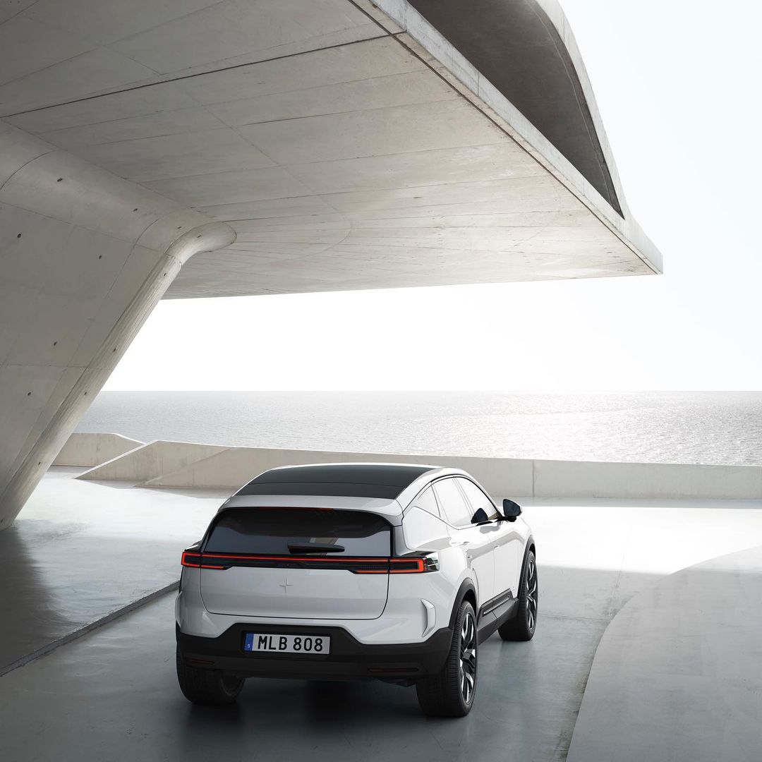 Rear view of a white Polestar SUV with a black roof parked under a concrete structure with the ocean in the background on a sunny day.
