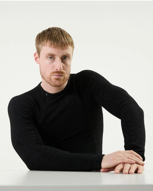 Studio shot of a fair-skinned adult man in a black long-sleeved textured shirt, with folded hands on a grey surface against a white backdrop.