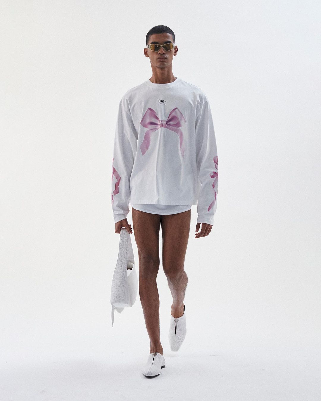 Fashion shot of a man with sunglasses and a long-sleeve t-shirt with a bow print, walking in front of a plain white backdrop holding a bag.