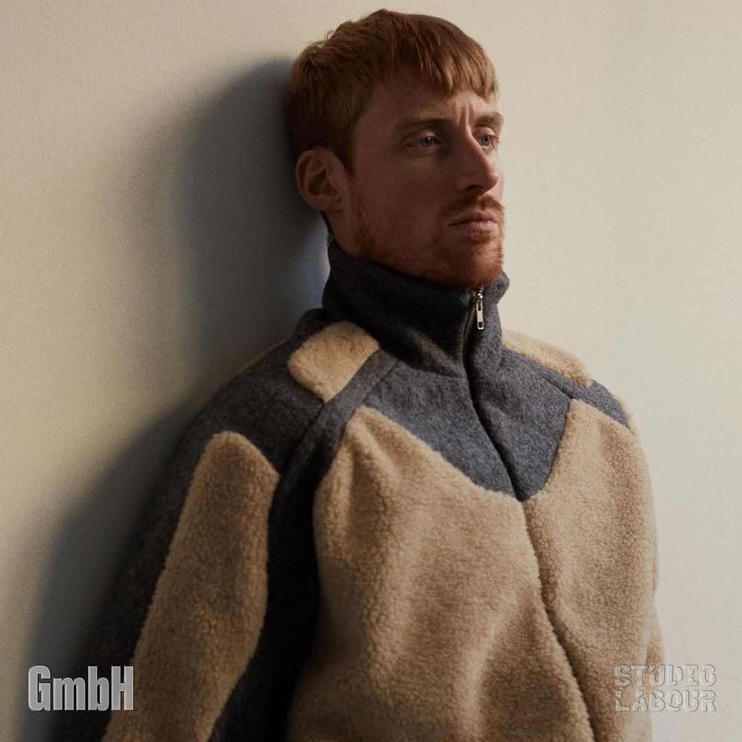 Man with red hair and beige jacket poses indoors against a wall. He is standing and facing right, eyes looking forward. Studio lighting.