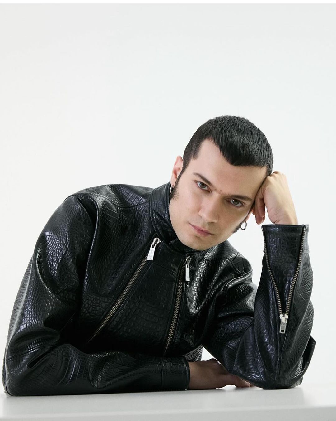 Adult male wearing a black faux-leather jacket with silver zippers rests his head on his hand while sitting against a white backdrop.