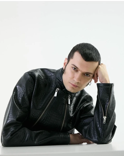 Adult male wearing a black faux-leather jacket with silver zippers rests his head on his hand while sitting against a white backdrop.