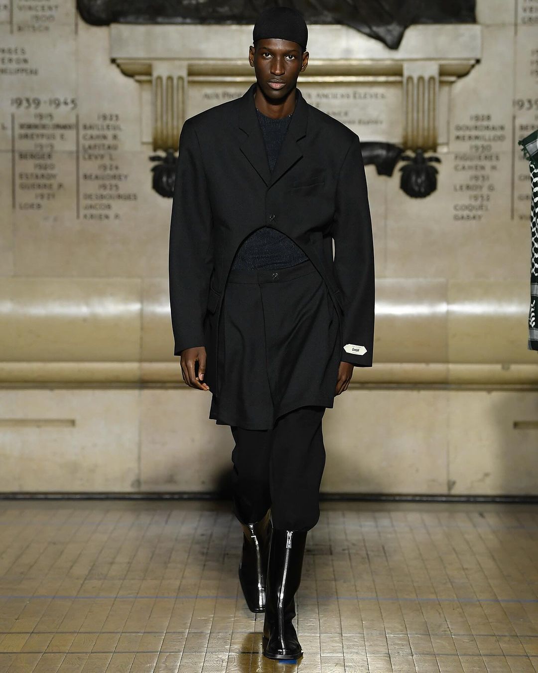 A dark-skinned male fashion model in a black suit and boots walks the runway, wearing a black beanie in an indoor setting.