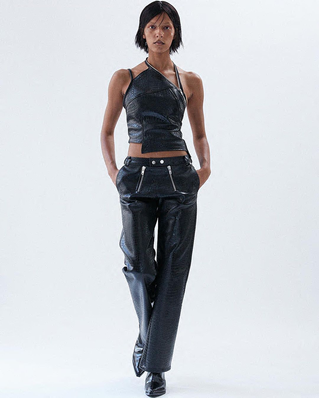Fashion model wears a dark, shiny, two-piece halter top and pantsuit with zipper details, posing against a bright white backdrop in a studio shot.