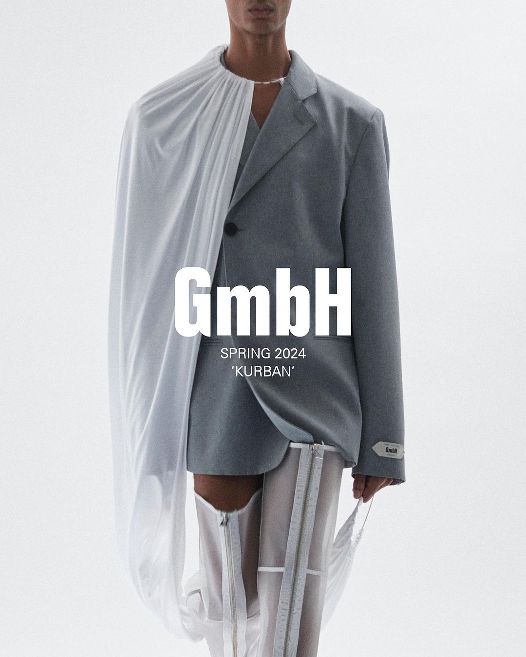 Runway fashion shot: a slender, light-skinned model showcases a gray suit paired with a white fabric drape and translucent boots in a studio.