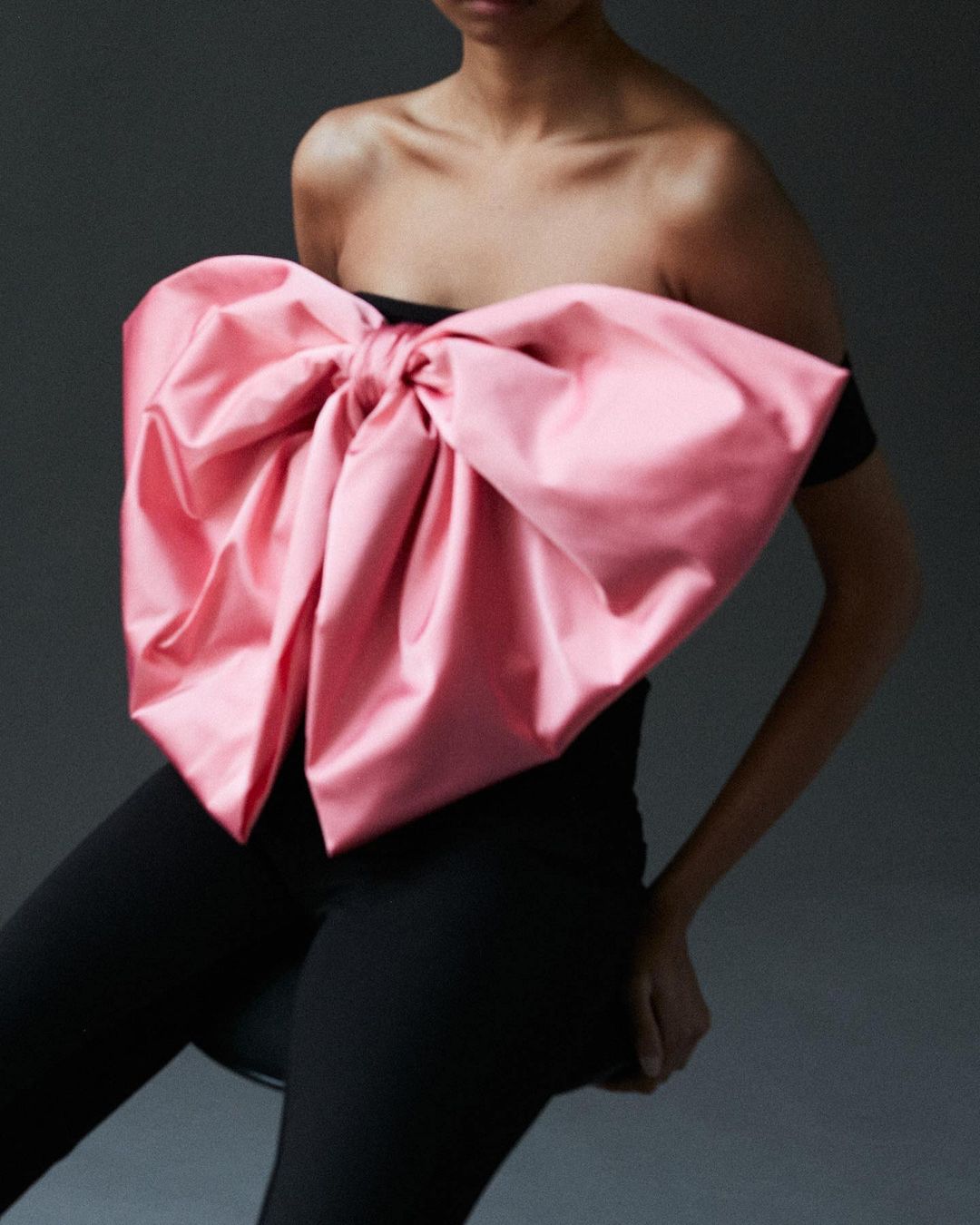 Fashion shot of a woman wearing black pants and a black top with a large pink bow, posing in front of a grey background. 