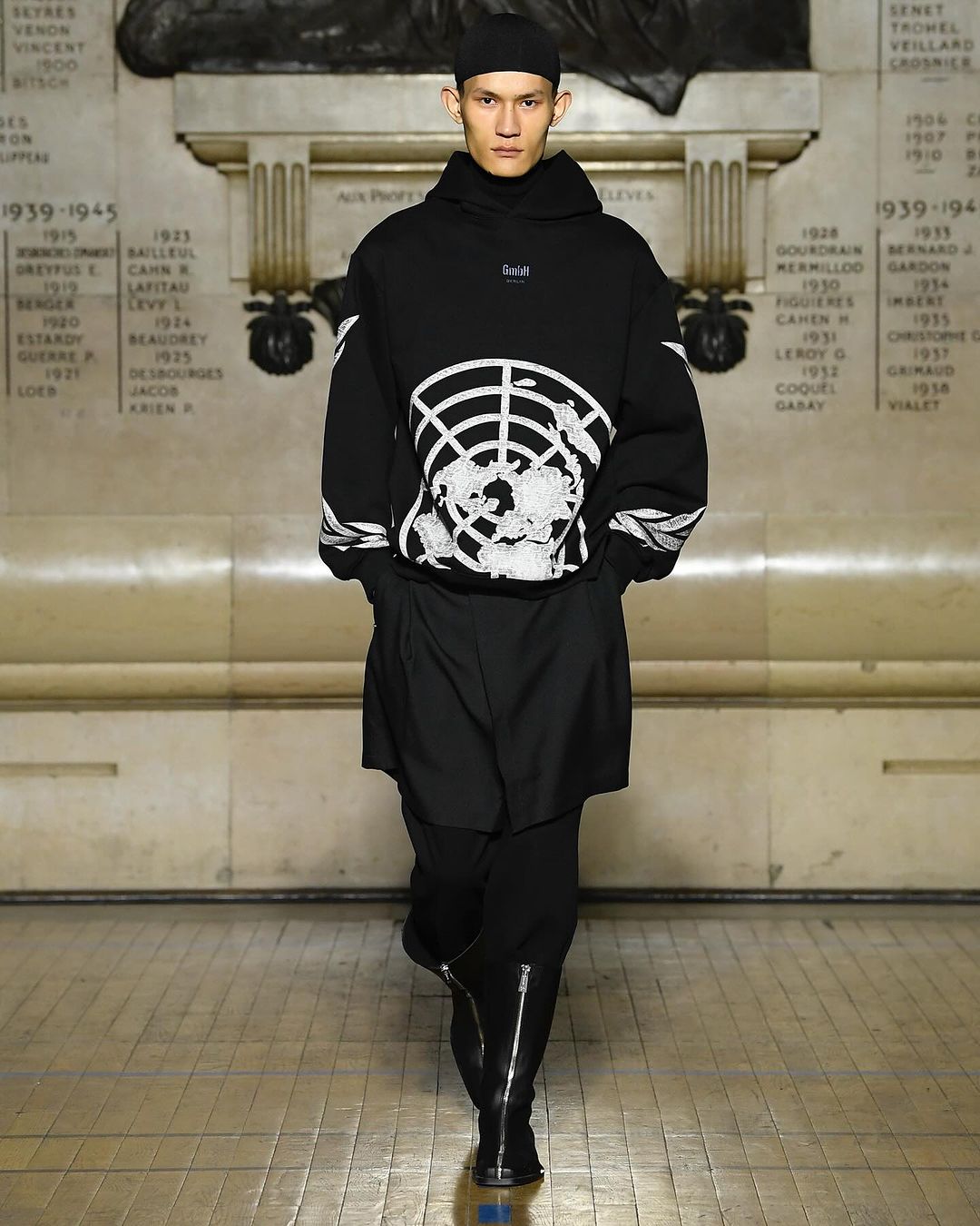 Asian male fashion model wearing a black hoodie and shorts walks a runway with tan stone walls and flooring in a fashion show.