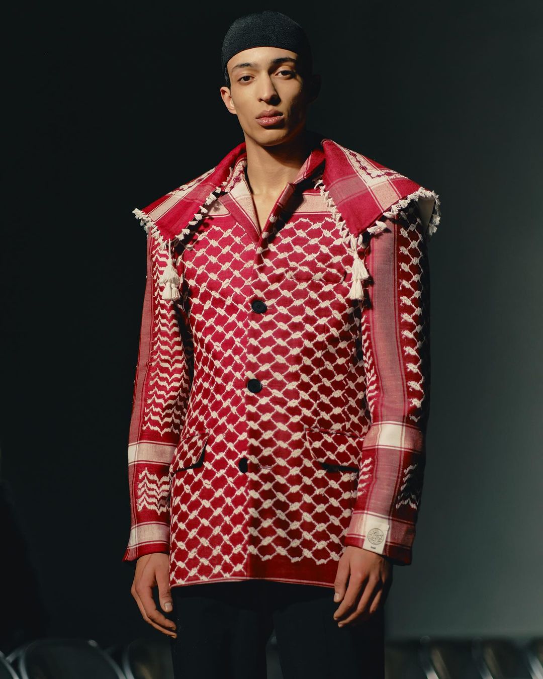 Young adult poses in a studio wearing a red patterned jacket with black pants. The jacket features draped tassels and buttons.