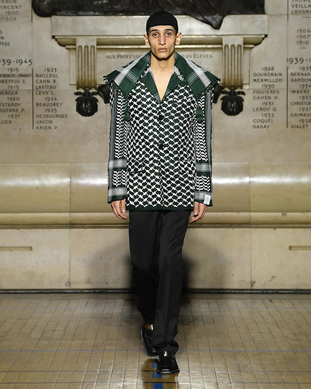 A male model walks the runway wearing a patterned jacket, black pants and shoes, and a black beanie in a well-lit indoor setting.