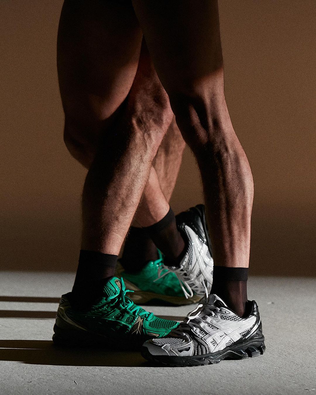 Two sets of legs wearing black socks and sneakers in neutral studio lighting. One pair of sneakers is green, and the other is silver.