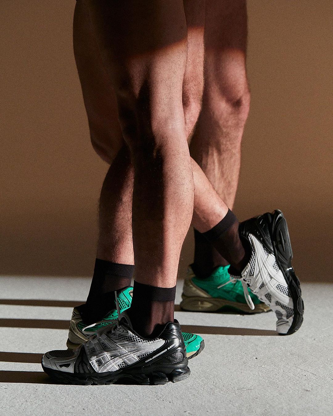Close-up of two adult male models standing indoors, wearing black socks and green and grey running shoes against a beige backdrop.