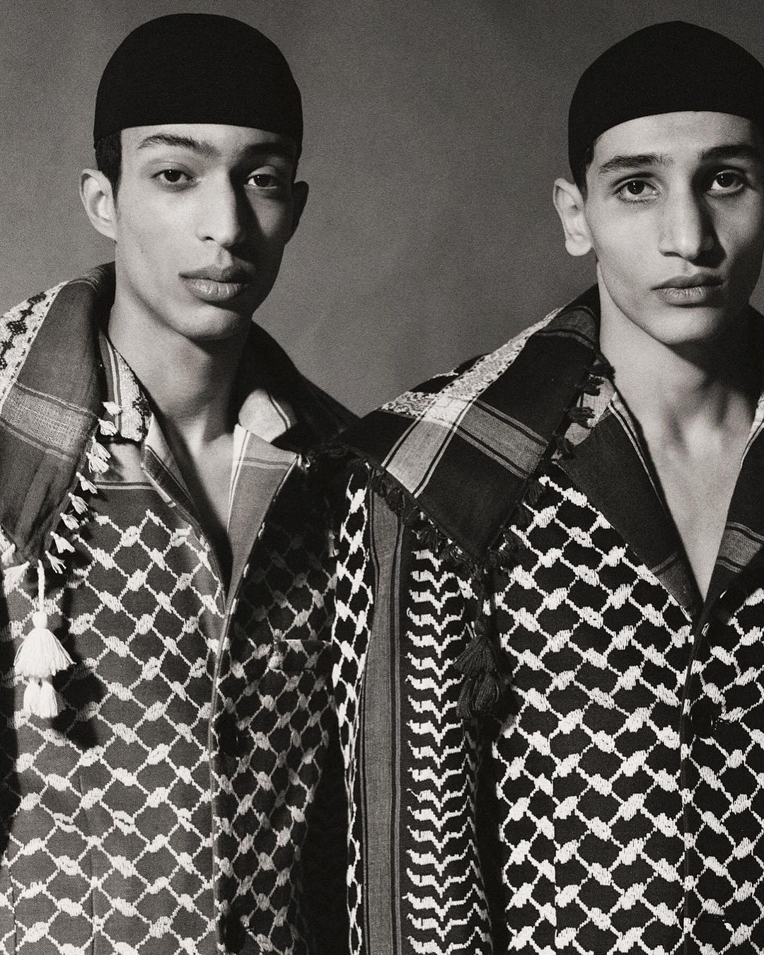 Two adult males wearing matching kufiyas and black caps pose indoors against a neutral backdrop, showcasing fashion and cultural attire.