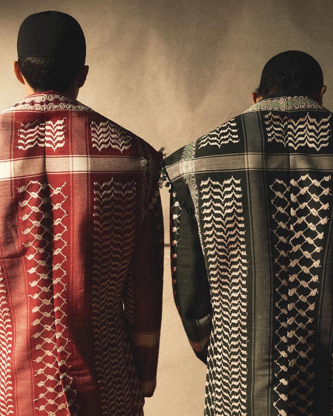 Two adults with their backs to the camera wearing matching keffiyeh and black taqiyah against a neutral background in a studio setting.