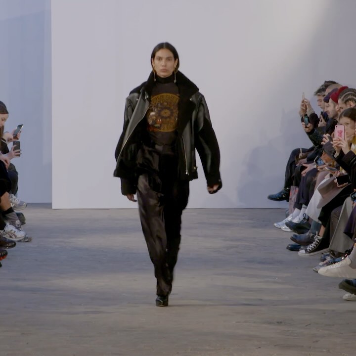 Runway fashion show featuring a model wearing a black leather jacket and shiny black pants walking toward the camera on a grey runway.