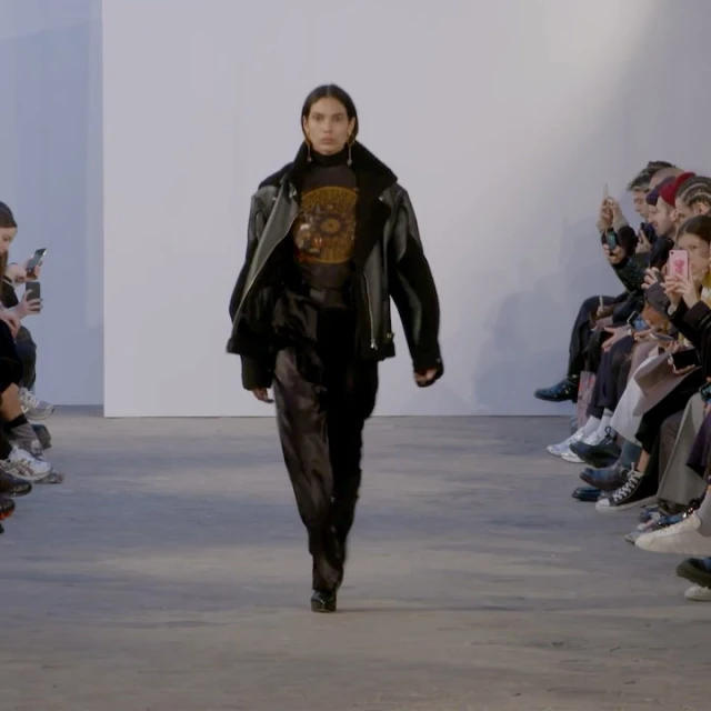 Runway fashion show featuring a model wearing a black leather jacket and shiny black pants walking toward the camera on a grey runway.