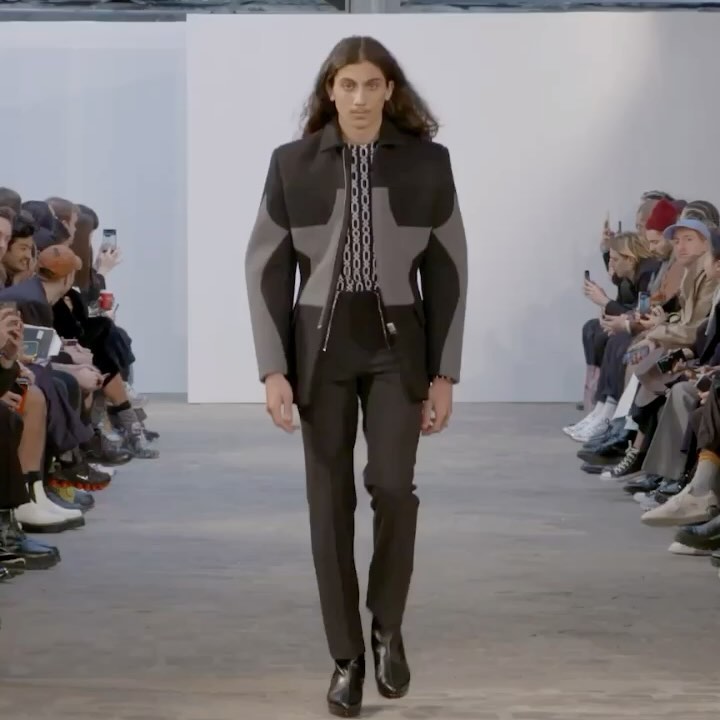 Runway fashion show featuring a male model in a grey and black suit, walking towards the camera, with an audience blurred in background.