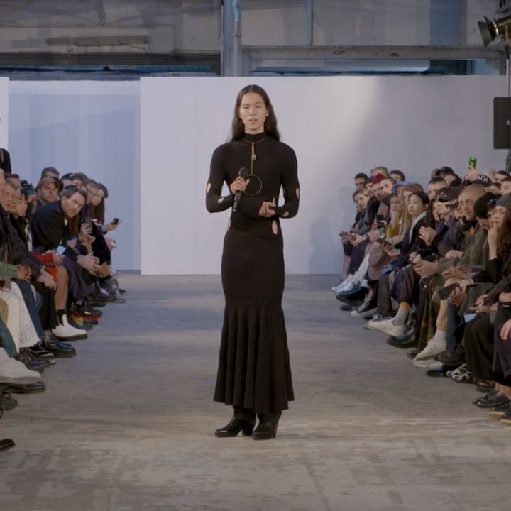 Runway show: a woman in a black dress walks a concrete runway, surrounded by a seated audience, in a brightly lit indoor space.