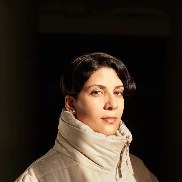 Portrait of an adult woman with short dark hair, wearing a light beige puffer jacket, posing indoors with a dark, shadowy background.