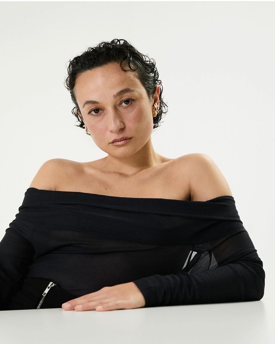 Close-up shot of a woman with short, dark hair, wearing a black, off-the-shoulder long-sleeve top against a bright white background.