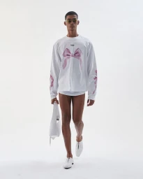 Model wearing white long-sleeve shirt with pink bow graphic, holding white handbag against white background