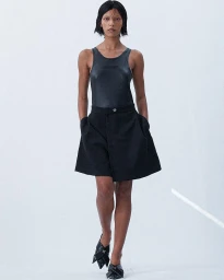 Model with dark short hair wearing a black sleeveless top and dark flared shorts against a plain white background.