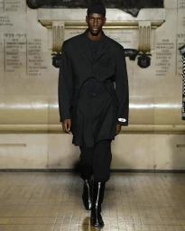 Male model in black suit jacket and trousers walking down runway with memorial wall background