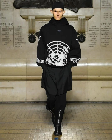 Model in black hoodie with white graphic print walking down a runway in a hall with marble walls