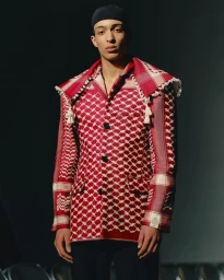 Model wearing a red and white patterned keffiyeh blazer and a black cap against a dark moody studio background.