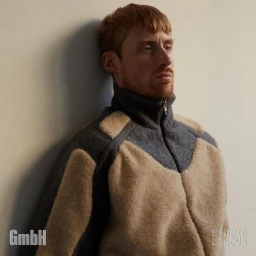 Red-haired man leaning against a light-coloured wall wearing a fleece jacket with chevron paneling.