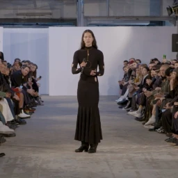Model with long dark hair wearing a black long-sleeved dress with cutout details standing on a runway