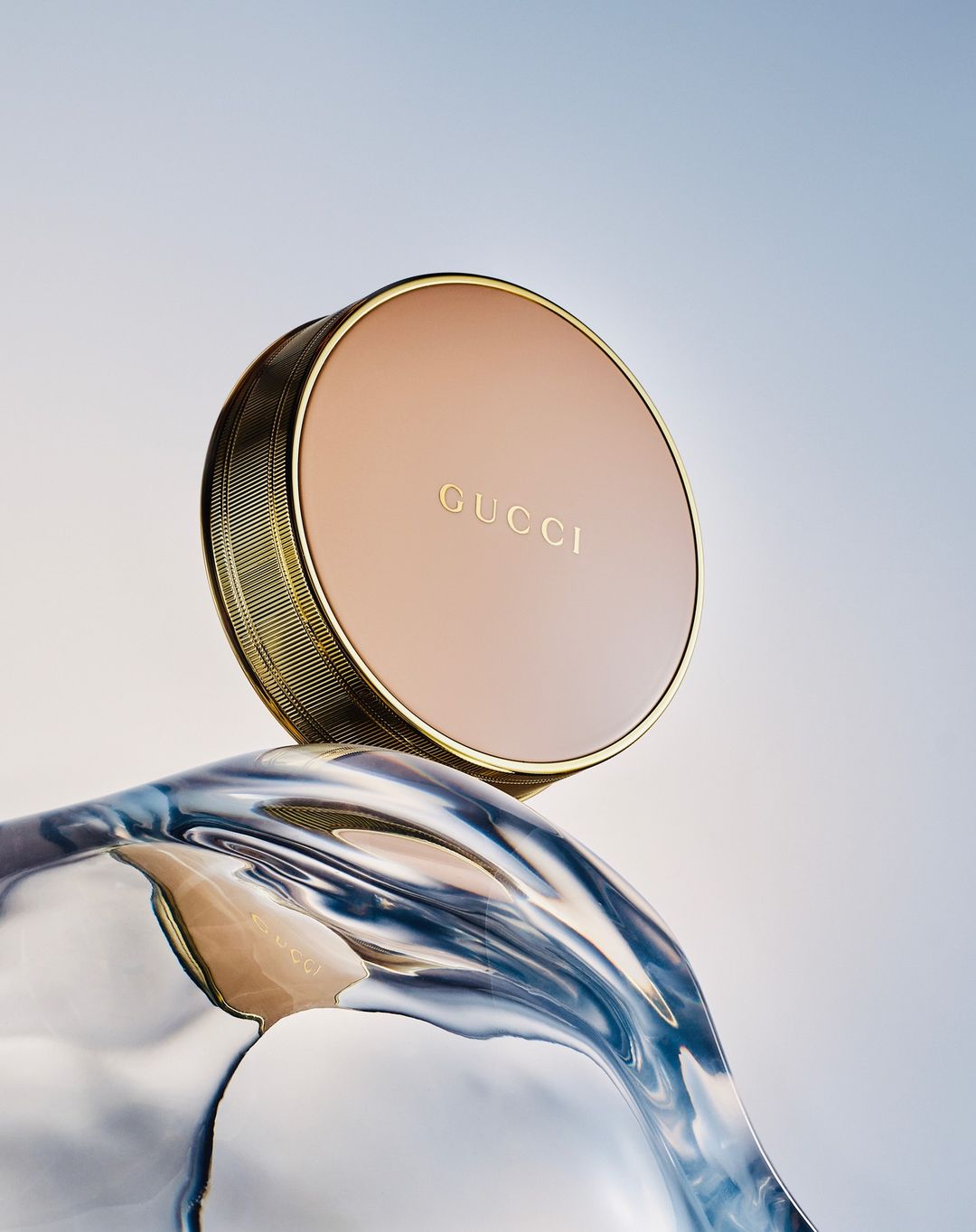 Close-up studio shot of a gold Gucci compact on swirling translucent blue material against a light blue gradient background.