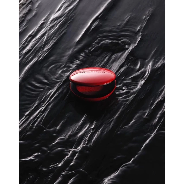 Red powder compact from WESTMAN ATELIER on black textured background in beauty advertising layout with simple elegance and studio lighting.