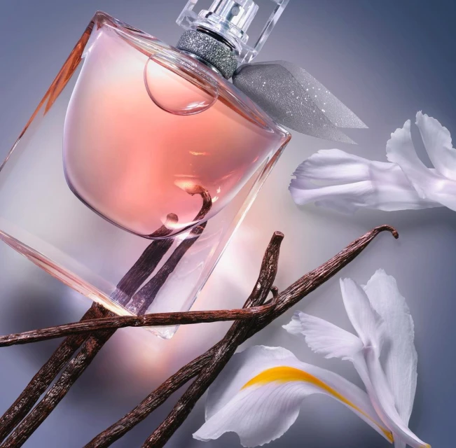 Studio shot of a clear glass perfume bottle with pale pink fragrance, decorated with a silver bow, vanilla beans and white flower petals.