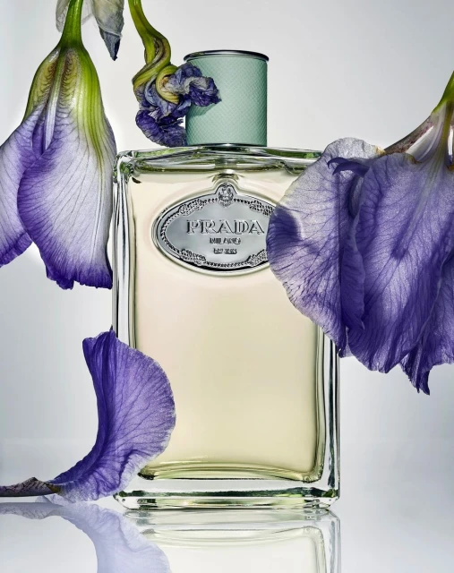 Close-up studio shot of a Prada perfume bottle surrounded by violet iris flowers, set against a soft white background with reflective surface.