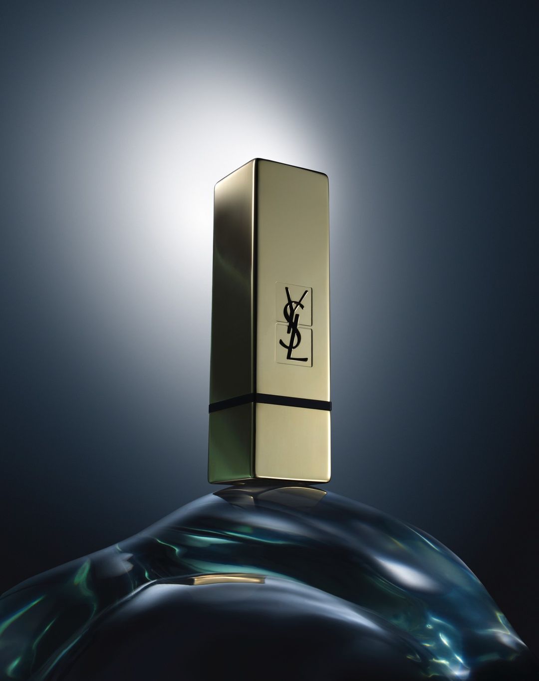 Gold YSL lipstick tube stands on a blue, reflective, curved surface against a grey-blue background with central white spot lighting.