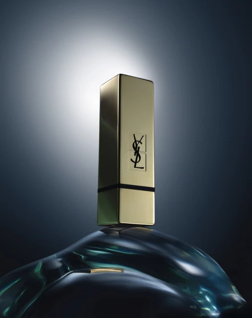 Gold YSL lipstick tube stands on a blue, reflective, curved surface against a grey-blue background with central white spot lighting.