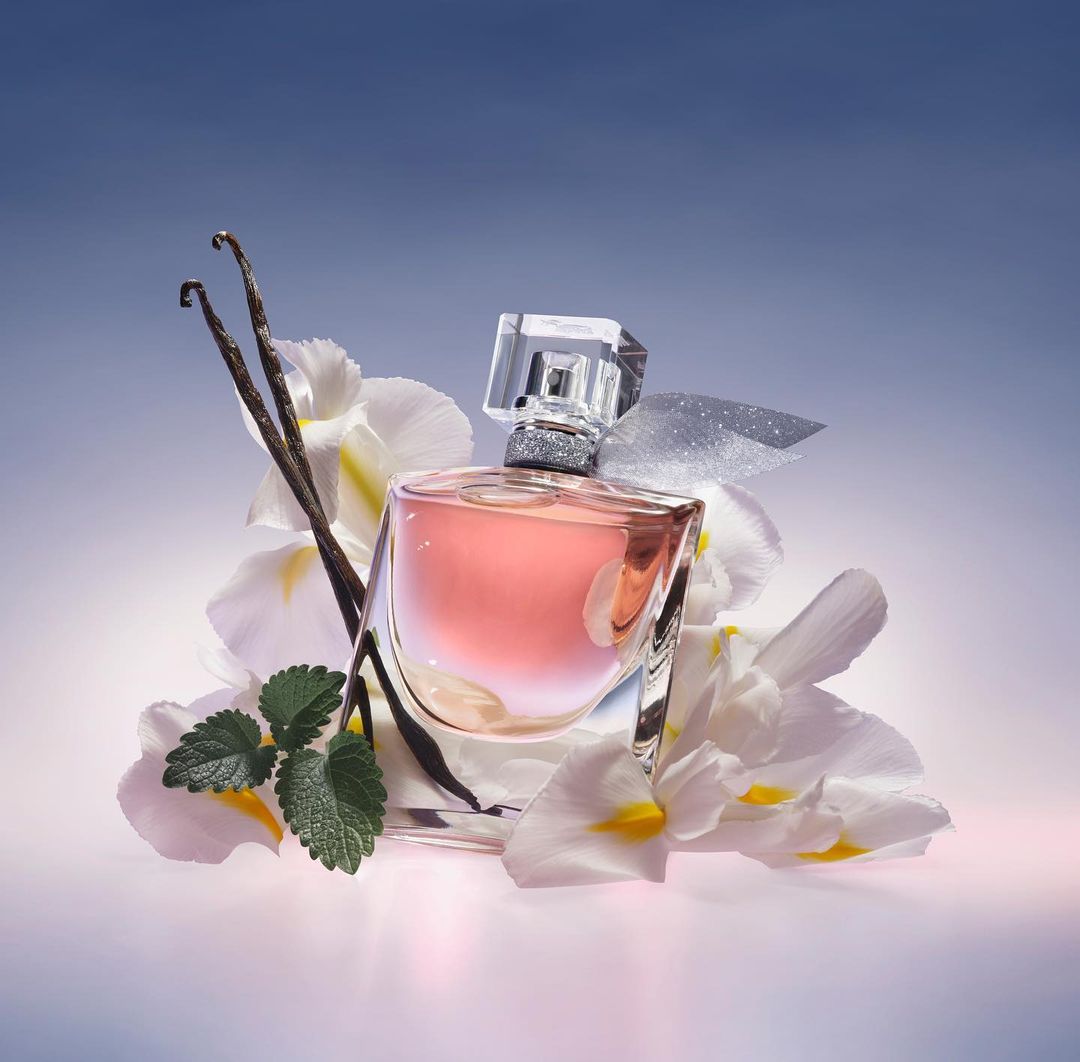 Elegant still life captures a perfume bottle nestled amidst delicate white flowers, vanilla beans, and green leaves against a soft blue backdrop.