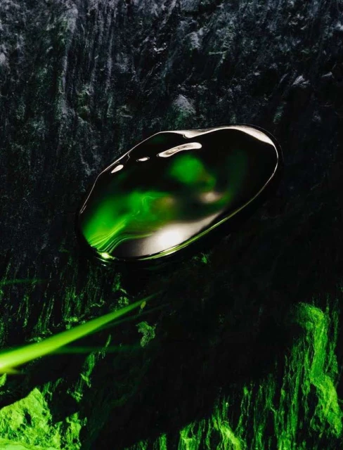 Dark textured surface features a droplet or pool of emerald-green liquid illuminated by a bright light source.