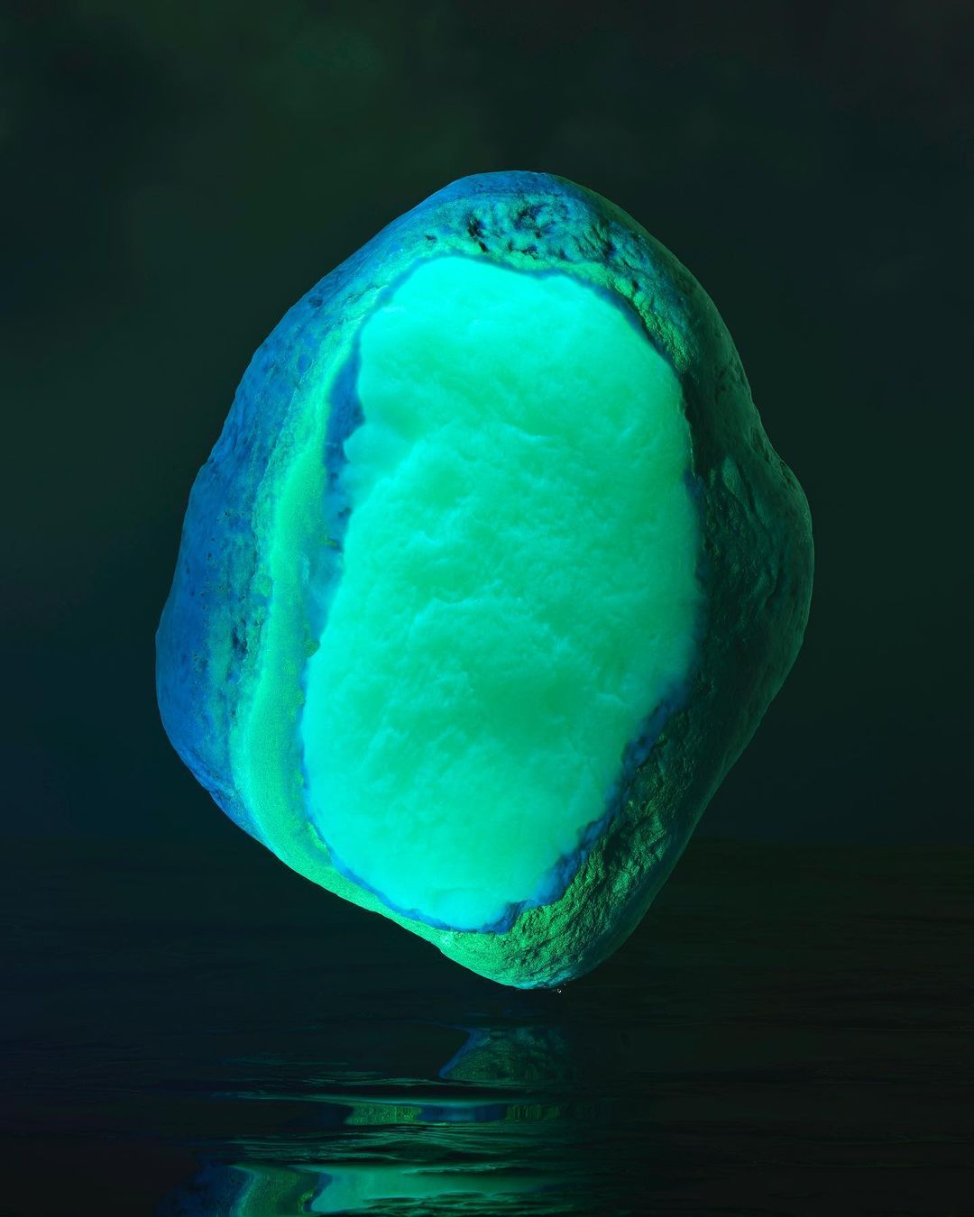 A green and blue abstract rock-like object levitates above a reflective dark surface, creating a visually striking and otherworldly image.