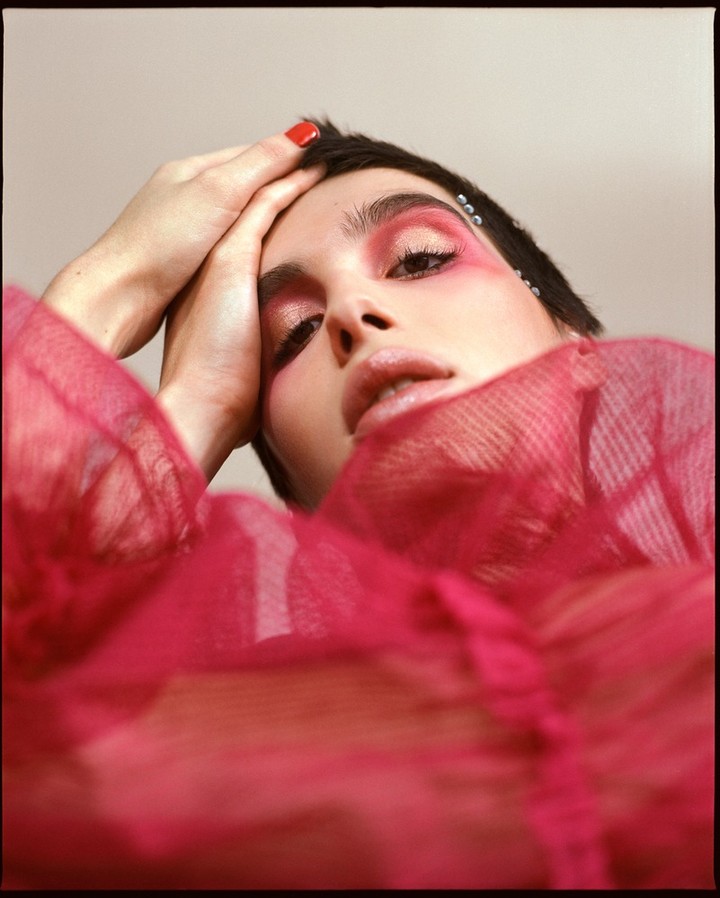 Close-up portrait of a woman with short black hair and red makeup, posing with her hands near her head, wearing a pink top.