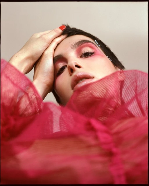 Close-up portrait of a woman with short black hair and red makeup, posing with her hands near her head, wearing a pink top.