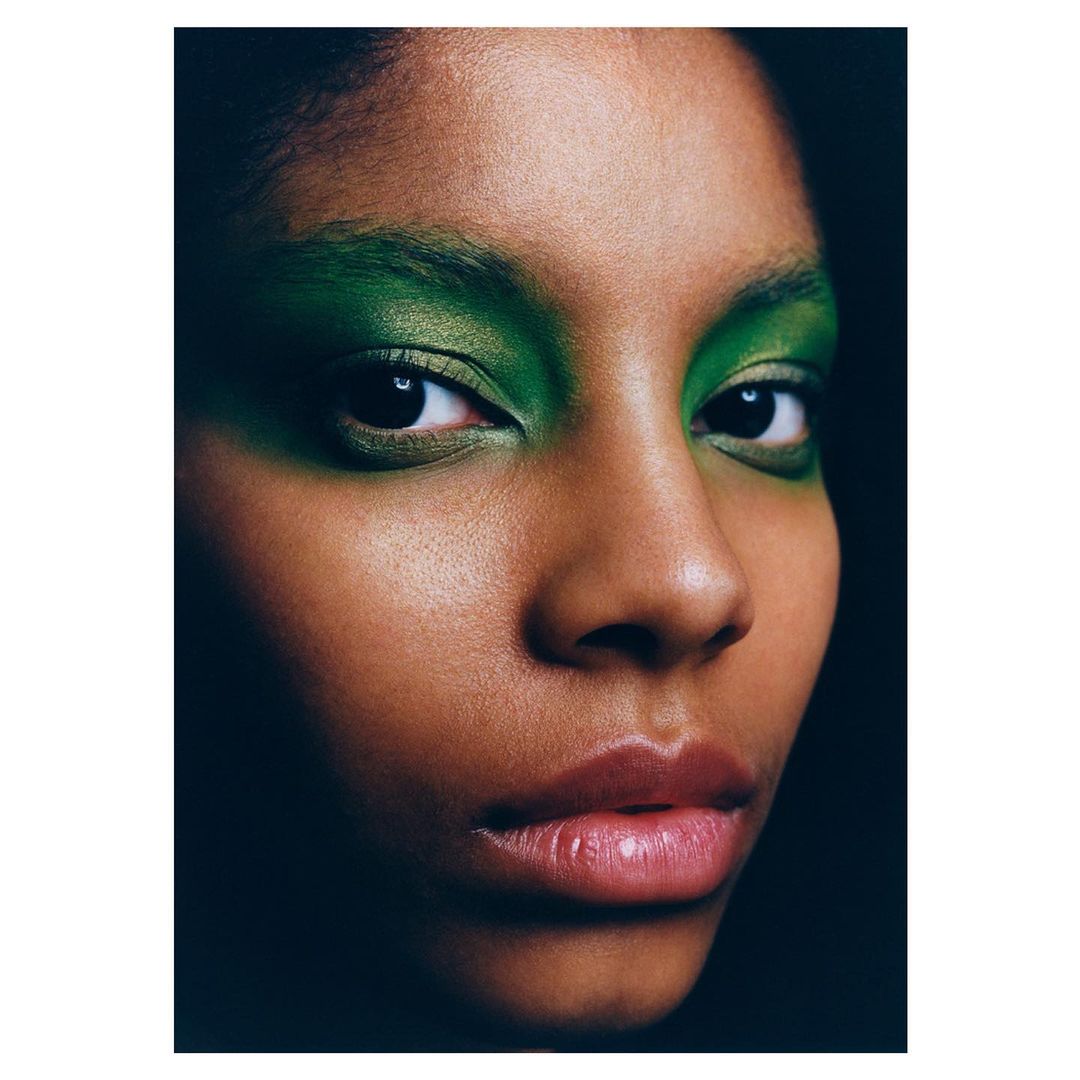 Close-up portrait of an African-American woman with green eyeshadow and glossy lips against a dark background in a studio shot.