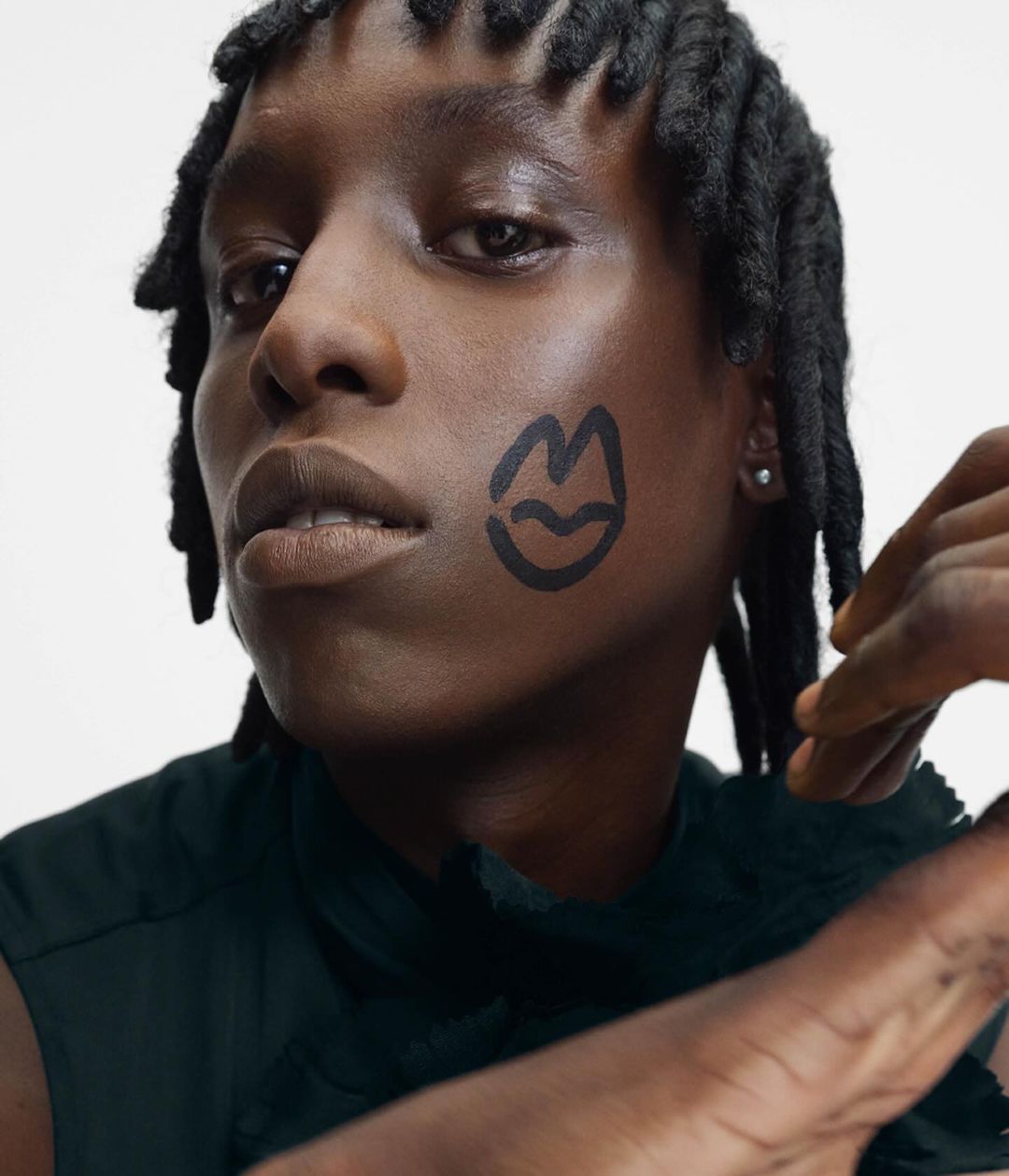 Close-up portrait of a Black adult with locs and an illustrative mouth drawn on their cheek, set against a plain white background.
